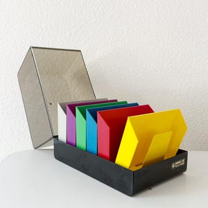 May include: A black organizer box with a clear lid, holding colorful, angled file holders. The file holders are in various colors: white, purple, green, blue, red, and yellow. The box has a label on the side.