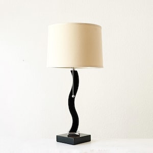 May include: A table lamp featuring a cylindrical beige fabric shade and a black, S-shaped curved stand. The base is a solid black square block, and a silver pull chain hangs from the center of the lamp socket beneath the shade.