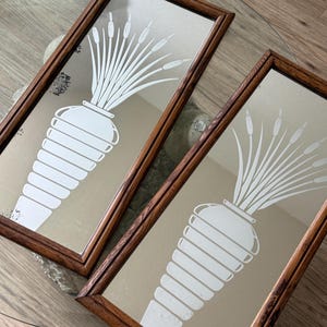 May include: Two antique mirrors with wooden frames, each featuring a white etched design of a vase with reeds. The mirrors are displayed on a glass table.