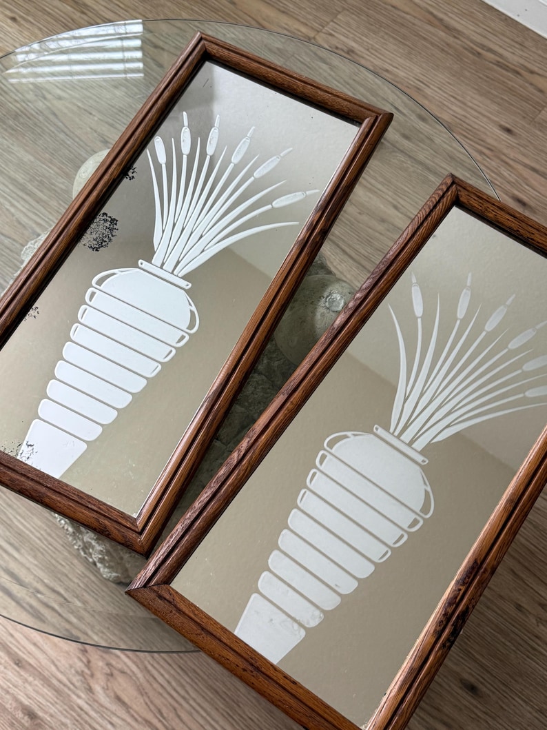 May include: Two antique mirrors with wooden frames, each featuring a white etched design of a vase with reeds. The mirrors are displayed on a glass table.