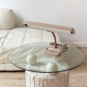 May include: A vintage, brown,  adjustable desk lamp with a white shade sits on a round glass table with a white stone base. Three white spheres are on the table.