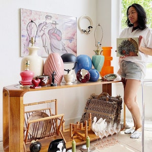 May include: A wooden console table with a variety of vintage decor items, including lamps, vases, and figurines. The table has a curved design and a light wood finish. There is a wicker magazine rack and a black and white floral vase on the table. A woman is standing behind the table, holding a book.