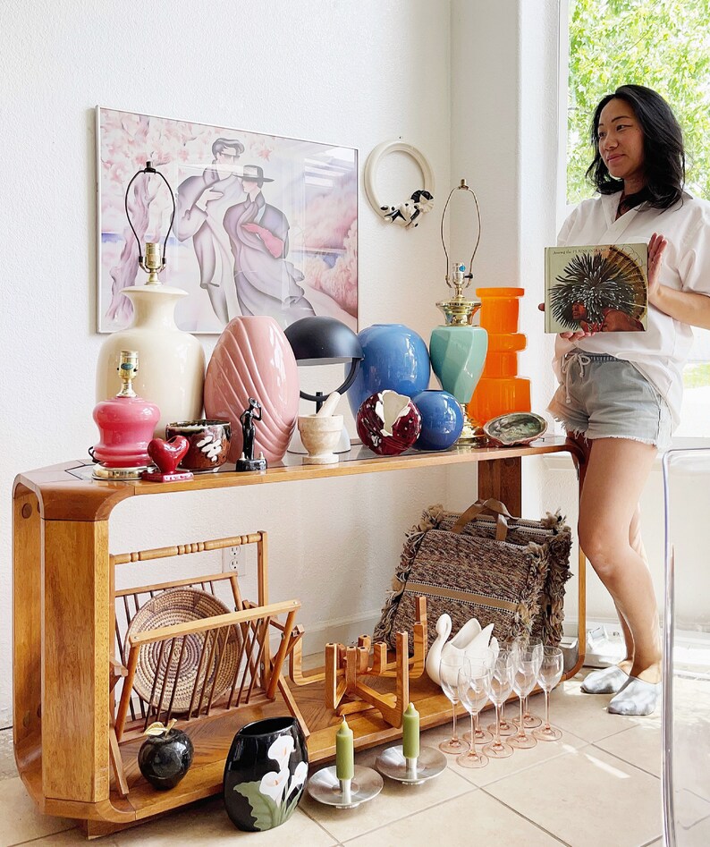 May include: A wooden console table with a variety of vintage decor items, including lamps, vases, and figurines. The table has a curved design and a light wood finish. There is a wicker magazine rack and a black and white floral vase on the table. A woman is standing behind the table, holding a book.