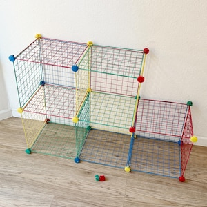 May include: A modular storage unit constructed from a grid of thin metal wires in red, green, yellow, and blue. The structure features five open cube compartments arranged in a stepped formation, connected by circular plastic joints in matching colors.