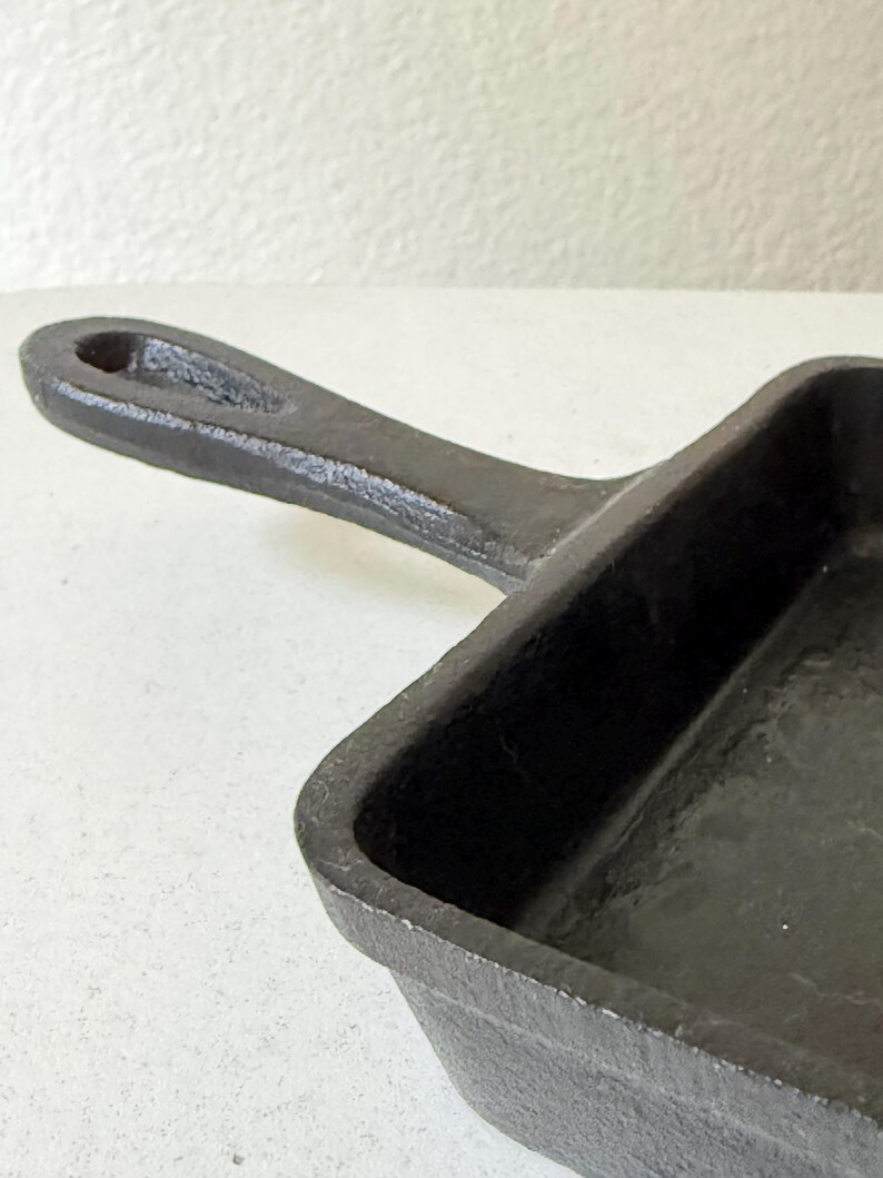 Puede incluir: Una sart&eacute;n de hierro fundido negro con una superficie de cocci&oacute;n rectangular y un asa. El asa tiene un agujero para colgar. La sart&eacute;n est&aacute; hecha de metal resistente y est&aacute; dise&ntilde;ada para cocinar.