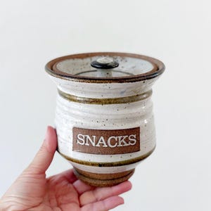 May include: White ceramic jar with a brown lid and a brown label that says "SNACKS".
