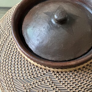 Op de afbeelding: Donkerbruine keramische pot met deksel, versierd met een ster en een knop. De pot staat op een basis geweven van natuurlijke vezels, waardoor een gestructureerd patroon ontstaat. Handwerk mogelijk.