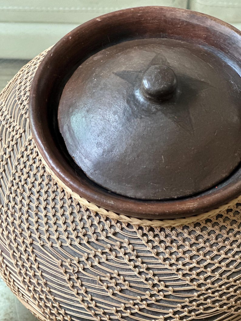 Op de afbeelding: Donkerbruine keramische pot met deksel, versierd met een ster en een knop. De pot staat op een basis geweven van natuurlijke vezels, waardoor een gestructureerd patroon ontstaat. Handwerk mogelijk.
