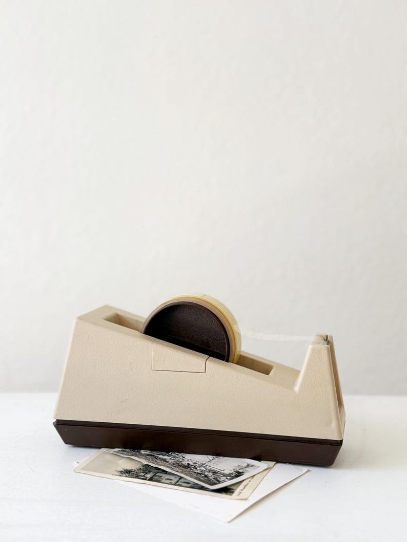 Puede incluir: Un dispensador de cinta adhesiva beige y marr&oacute;n con un rollo de cinta transparente. El dispensador tiene una base rectangular y una parte superior inclinada. Una pila de fotograf&iacute;as antiguas en blanco y negro descansa frente al dispensador.