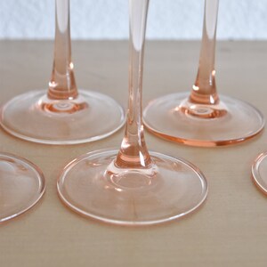 May include: Four pink wine glasses with clear stems and bases. The glasses are arranged in a row on a light wood surface.