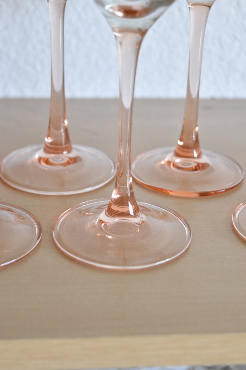 May include: Four pink wine glasses with clear stems and bases. The glasses are arranged in a row on a light wood surface.