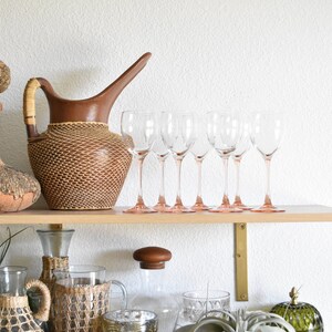 May include: A wooden shelf with a variety of decorative items, including a brown woven pitcher, clear glass wine glasses, a glass bottle with a wooden stopper, a woven glass bottle, a woven glass jar, two white ceramic birds, and a green glass jar with a decorative knob.
