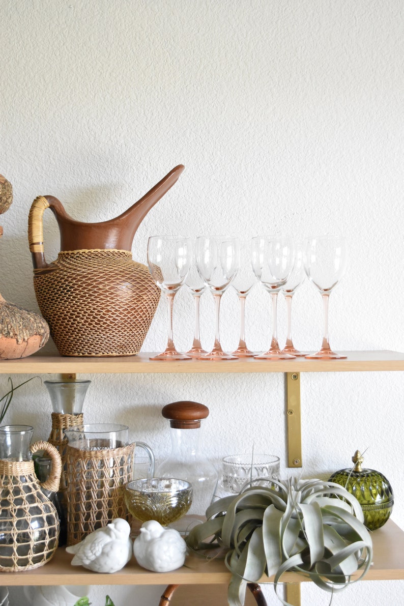 May include: A wooden shelf with a variety of decorative items, including a brown woven pitcher, clear glass wine glasses, a glass bottle with a wooden stopper, a woven glass bottle, a woven glass jar, two white ceramic birds, and a green glass jar with a decorative knob.