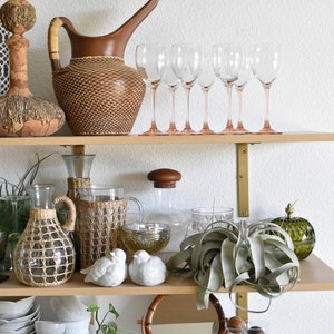 May include: A wooden shelf with various decorative items, including a wicker pitcher, glass decanters, and a green glass vase. There are also several clear glass wine glasses, a potted air plant, and a collection of white ceramic birds.