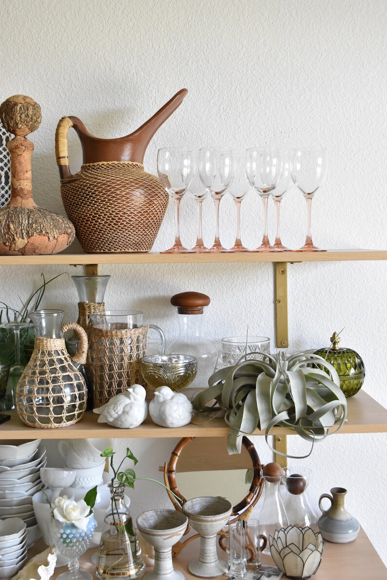 May include: A wooden shelf with various decorative items, including a wicker pitcher, glass decanters, and a green glass vase. There are also several clear glass wine glasses, a potted air plant, and a collection of white ceramic birds.