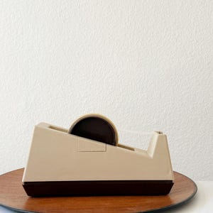 Puede incluir: Un dispensador de cinta adhesiva beige y marr&oacute;n con un rollo de cinta. El dispensador est&aacute; sobre un plato de madera. El fondo es una pared blanca.