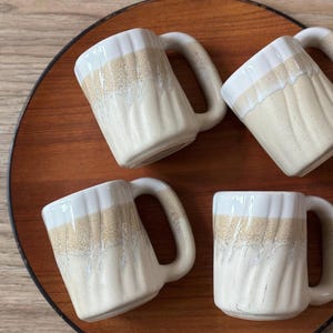 May include: Set of four white ceramic mugs with a brown glaze. The mugs have a textured, wavy design and are arranged on a wooden tray.