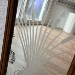 May include: A wooden framed mirror with a white etched design of a plant with tall leaves. The mirror is reflecting a wooden floor and a blurry view of a room.