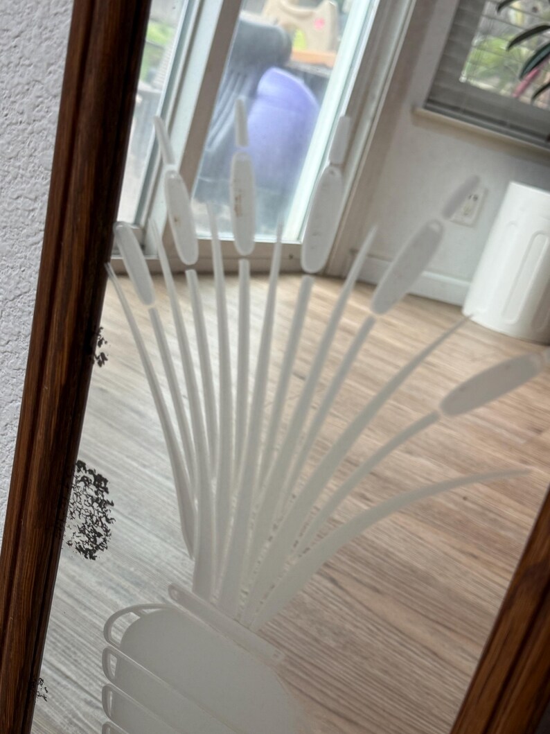 May include: A wooden framed mirror with a white etched design of a plant with tall leaves. The mirror is reflecting a wooden floor and a blurry view of a room.