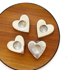 May include: Four silver heart-shaped candle holders arranged on a round wooden tray with a black trim. Each holder has a circular indentation for a candle. The tray has a warm, natural wood grain.