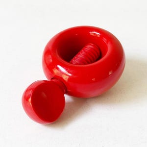 May include: A bright red nutcracker with a round body and a smaller, spherical handle. The interior features a ridged, cylindrical mechanism for cracking nuts. The nutcracker is made of a glossy material.