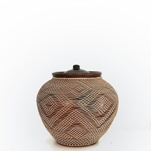Op de afbeelding: Een bruine keramische pot met een geweven, lichtbruin roosterontwerp. De pot heeft een donkerbruine rand en een bijpassende deksel. De pot is rond met een licht bolvormige vorm. De achtergrond is wit.