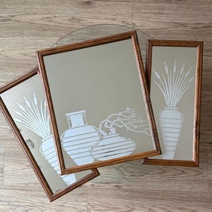 May include: Three framed mirrors with a beige background and white stylized illustrations of vases and reeds. The mirrors are in wooden frames with a dark brown finish.