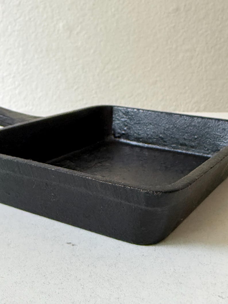Puede incluir: Primer plano de una peque&ntilde;a sart&eacute;n cuadrada de hierro fundido negro con un asa. La sart&eacute;n tiene un exterior texturizado y un interior liso. La sart&eacute;n est&aacute; dise&ntilde;ada para cocinar y probablemente se utiliza para porciones individuales.