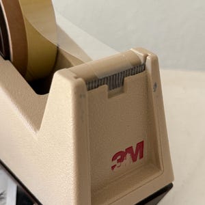 Puede incluir: Dispensador de cinta adhesiva 3M beige con un rollo de cinta. El dispensador tiene un logotipo rojo 3M en el lateral. La cinta es transparente y marr&oacute;n. El dispensador est&aacute; sobre una superficie blanca.
