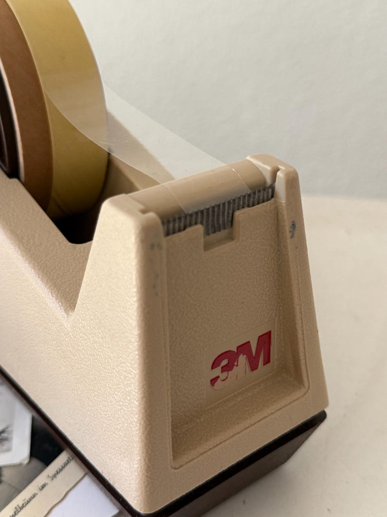 Puede incluir: Dispensador de cinta adhesiva 3M beige con un rollo de cinta. El dispensador tiene un logotipo rojo 3M en el lateral. La cinta es transparente y marr&oacute;n. El dispensador est&aacute; sobre una superficie blanca.