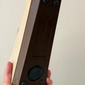 Puede incluir: Un objeto rectangular vintage con cuerpo crema y bordes marr&oacute;n oscuro. La base tiene cuatro peque&ntilde;os pies de goma y un componente negro circular. El objeto se sostiene en una mano.