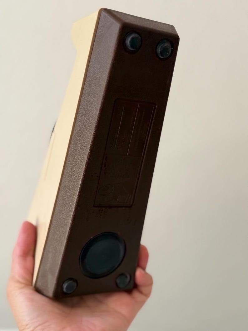 Puede incluir: Un objeto rectangular vintage con cuerpo crema y bordes marr&oacute;n oscuro. La base tiene cuatro peque&ntilde;os pies de goma y un componente negro circular. El objeto se sostiene en una mano.