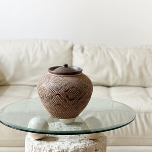 Op de afbeelding: Een bruine geweven pot met een deksel staat op een tafel met glazen blad. De pot heeft een textuurpatroon en een donkerbruin deksel en knop. De tafel wordt ondersteund door een witte, getextureerde basis. Op de achtergrond is een witte bank te zien.