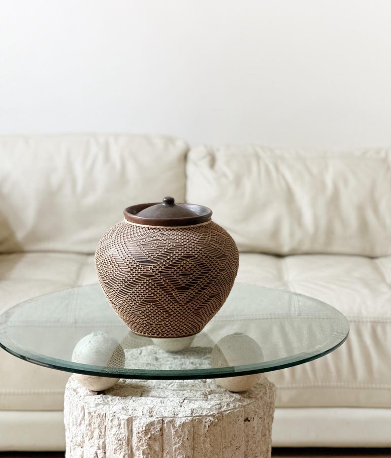 Op de afbeelding: Een bruine geweven pot met een deksel staat op een tafel met glazen blad. De pot heeft een textuurpatroon en een donkerbruin deksel en knop. De tafel wordt ondersteund door een witte, getextureerde basis. Op de achtergrond is een witte bank te zien.