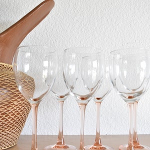 May include: Five clear wine glasses with pink stems stand on a light brown surface. The glasses are empty and appear to be made of glass.