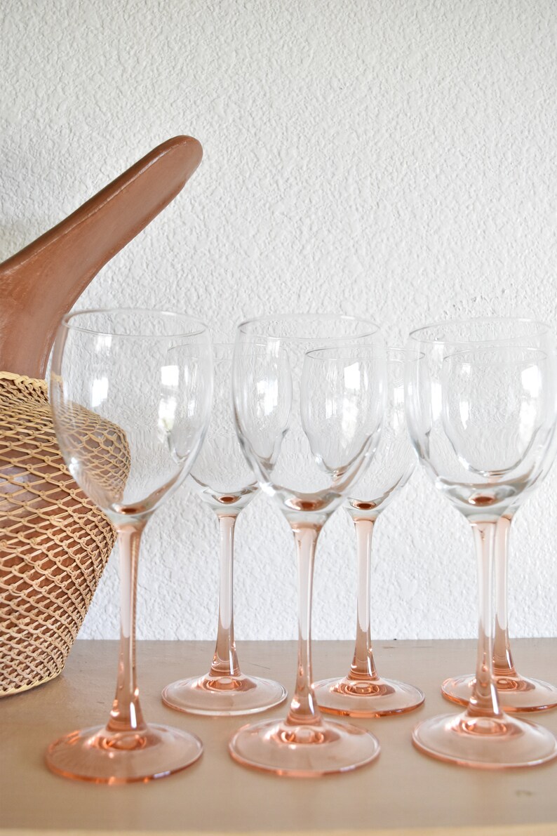 May include: Five clear wine glasses with pink stems stand on a light brown surface. The glasses are empty and appear to be made of glass.
