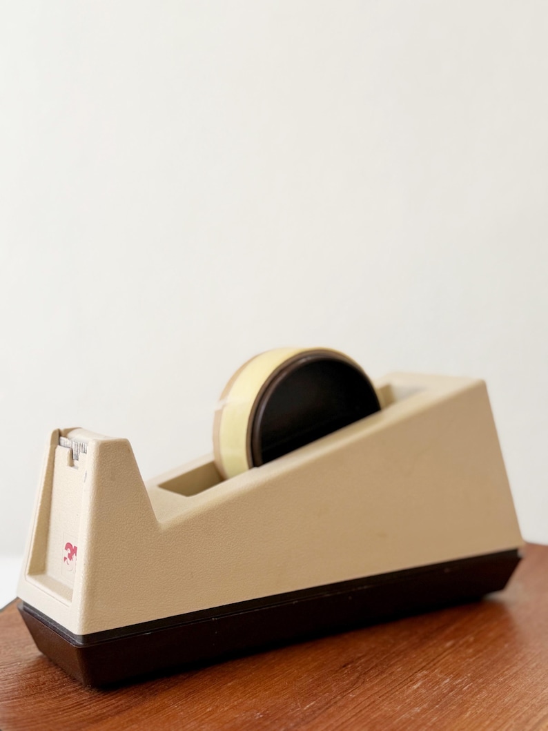 Puede incluir: Un dispensador de cinta adhesiva beige y marr&oacute;n con un rollo de cinta. El dispensador tiene una base rectangular y una parte superior inclinada con un borde de corte. El rollo de cinta es beige y negro. El dispensador est&aacute; sobre una superficie de madera.