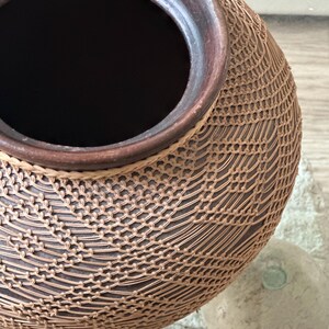 Op de afbeelding: Een handgemaakte, bruine keramische vaas met een geweven, netachtig patroon. De vaas heeft een donker interieur en een donkerbruine rand. De buitenkant heeft een lichtbruin, ingewikkeld ontwerp, waardoor een textuuroppervlak ontstaat. De vaas is een decoratief item.