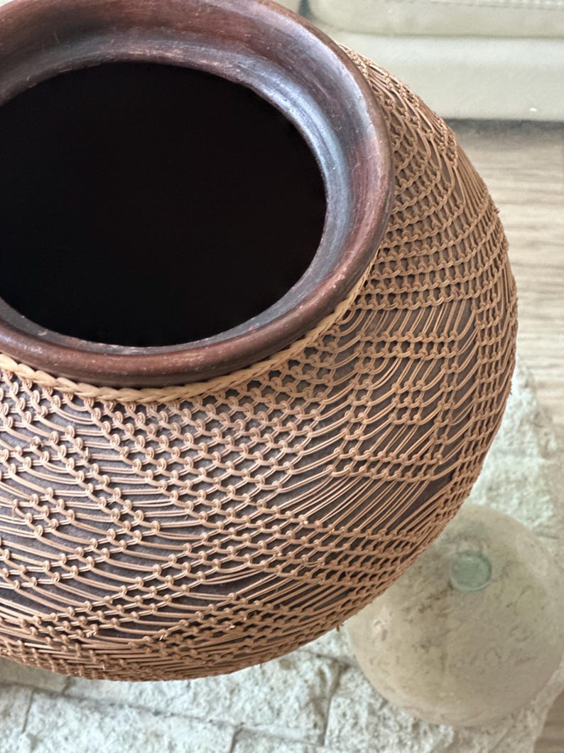 Op de afbeelding: Een handgemaakte, bruine keramische vaas met een geweven, netachtig patroon. De vaas heeft een donker interieur en een donkerbruine rand. De buitenkant heeft een lichtbruin, ingewikkeld ontwerp, waardoor een textuuroppervlak ontstaat. De vaas is een decoratief item.