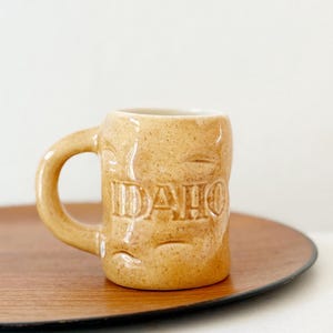 May include: A small, tan ceramic mug with the word "IDAHO" embossed on the front. The mug has a rounded handle and a speckled texture. It sits on a round, dark brown wooden tray.