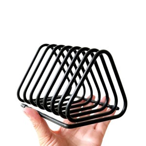 May include: Black metal napkin holder with a geometric design. The holder features a series of triangular shapes, providing a modern and minimalist aesthetic. The holder is designed to hold napkins or other paper items.