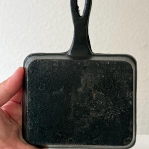 Puede incluir: Una peque&ntilde;a sart&eacute;n rectangular de hierro fundido negro con un asa. La sart&eacute;n tiene una superficie de cocci&oacute;n lisa y un lazo en el extremo del asa para colgarla. La sart&eacute;n est&aacute; sujeta por una mano.