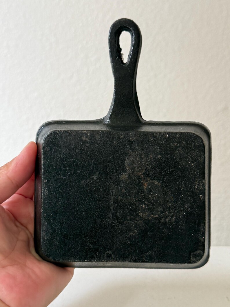 Puede incluir: Una peque&ntilde;a sart&eacute;n rectangular de hierro fundido negro con un asa. La sart&eacute;n tiene una superficie de cocci&oacute;n lisa y un lazo en el extremo del asa para colgarla. La sart&eacute;n est&aacute; sujeta por una mano.