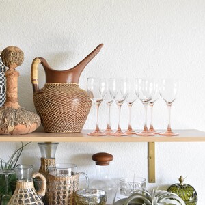 May include: A wooden shelf with a variety of decorative items, including a brown wicker pitcher, clear glass wine glasses with pink stems, a clear glass bottle with a wicker cover, a clear glass bottle with a wooden lid, a wicker bottle, a green glass bowl, a green glass jar, and a white ceramic bird figurine.