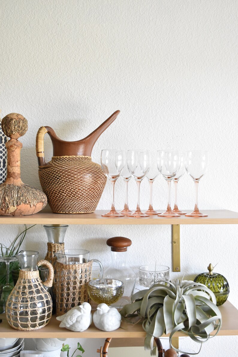 May include: A wooden shelf with a variety of decorative items, including a brown wicker pitcher, clear glass wine glasses with pink stems, a clear glass bottle with a wicker cover, a clear glass bottle with a wooden lid, a wicker bottle, a green glass bowl, a green glass jar, and a white ceramic bird figurine.