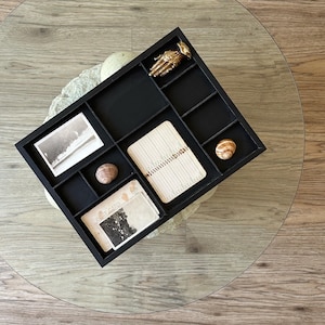 Könnte beinhalten: Ein schwarzes Holztablett mit Fächern, das eine alte Postkarte, ein kleines Notizbuch, eine goldfarbene Handfigur und zwei kleine runde Gegenstände enthält. Das Tablett befindet sich auf einer Glastischplatte.