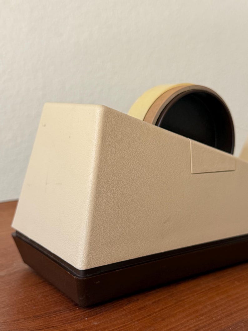Puede incluir: Un dispensador de cinta adhesiva beige y marr&oacute;n con un rollo de cinta. El dispensador tiene una superficie texturizada y una base marr&oacute;n oscuro. El rollo de cinta es visible en la parte superior.