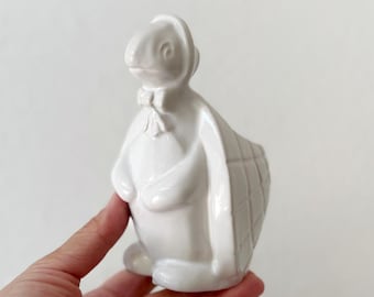 Op de afbeelding: Een witte keramische konijnenfiguur met een muts en strik. Het konijn heeft een mandachtig ontwerp op de rug. Het beeldje wordt in een hand gehouden tegen een witte achtergrond. Het konijn is een decoratief item.