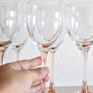 May include: Set of five clear glass wine glasses with pink stems and bases. The glasses are arranged in a row on a light surface.