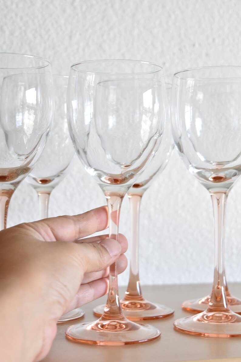 May include: Set of five clear glass wine glasses with pink stems and bases. The glasses are arranged in a row on a light surface.